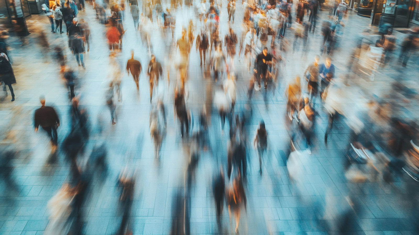 A motion-blurred overhead view of a crowded public walkway with people crossing a tiled plaza.