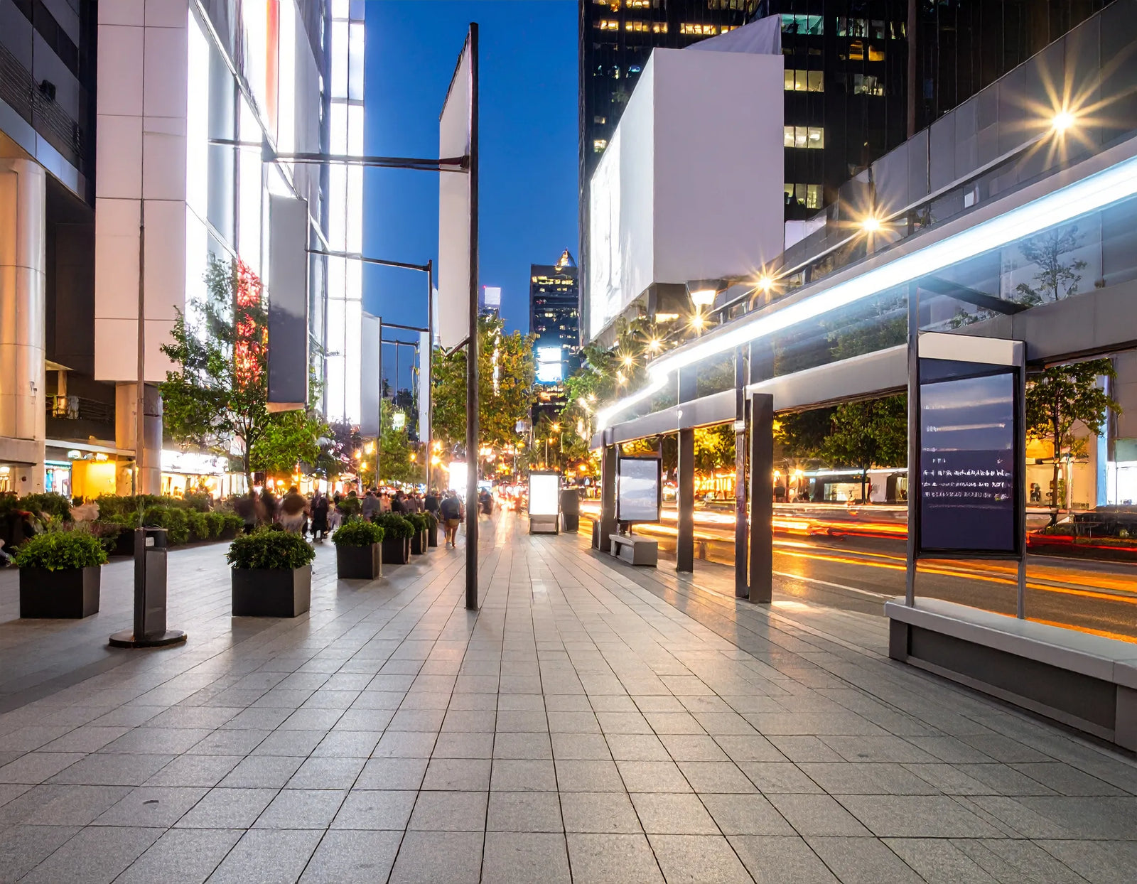 A wide urban plaza at dusk with a tiled walkway, lit storefronts and light trails.