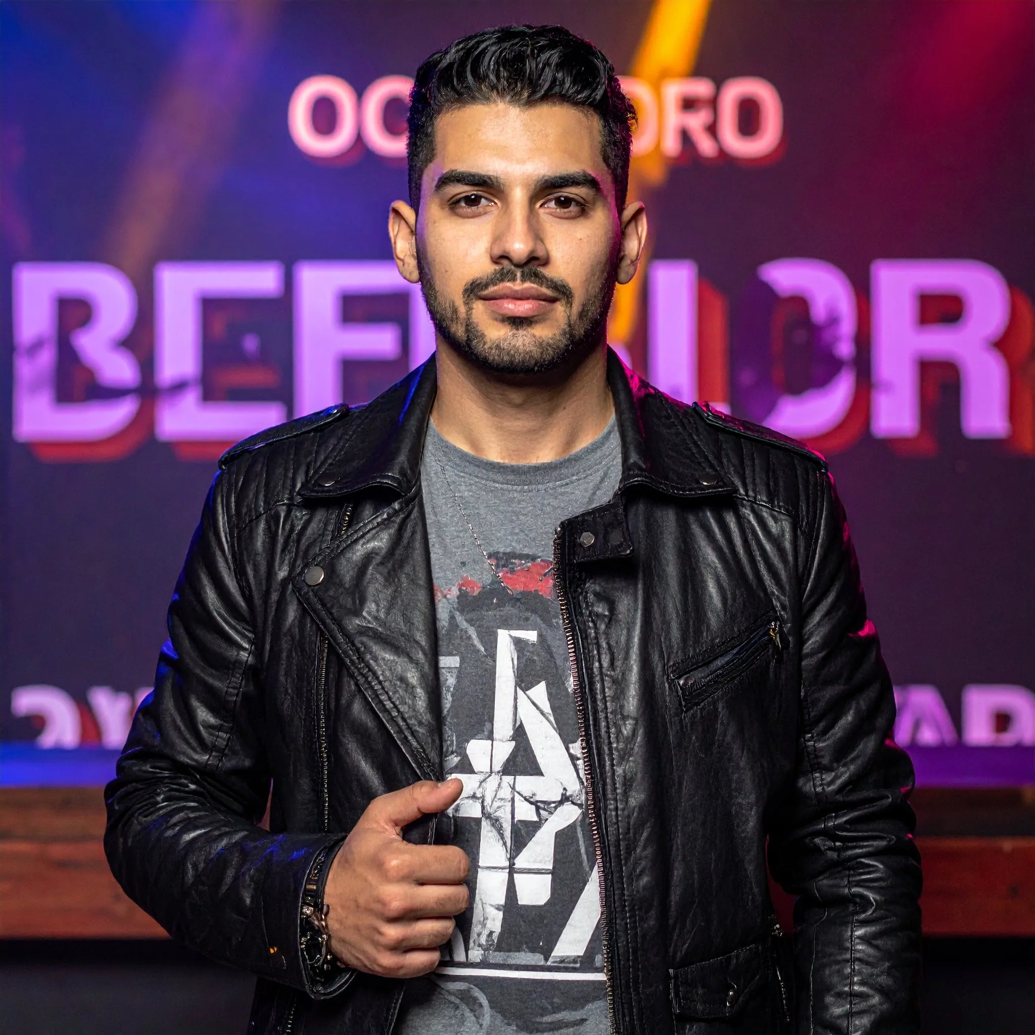 A man wearing a black leather jacket over a gray graphic t-shirt, standing in front of a purple and orange stage backdrop with the word 'BEFORE' visible.