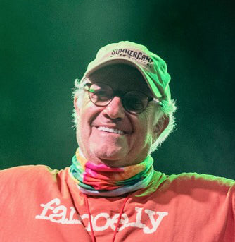 Person wearing a green cap and colorful scarf with a red shirt featuring white text, against a dark background.